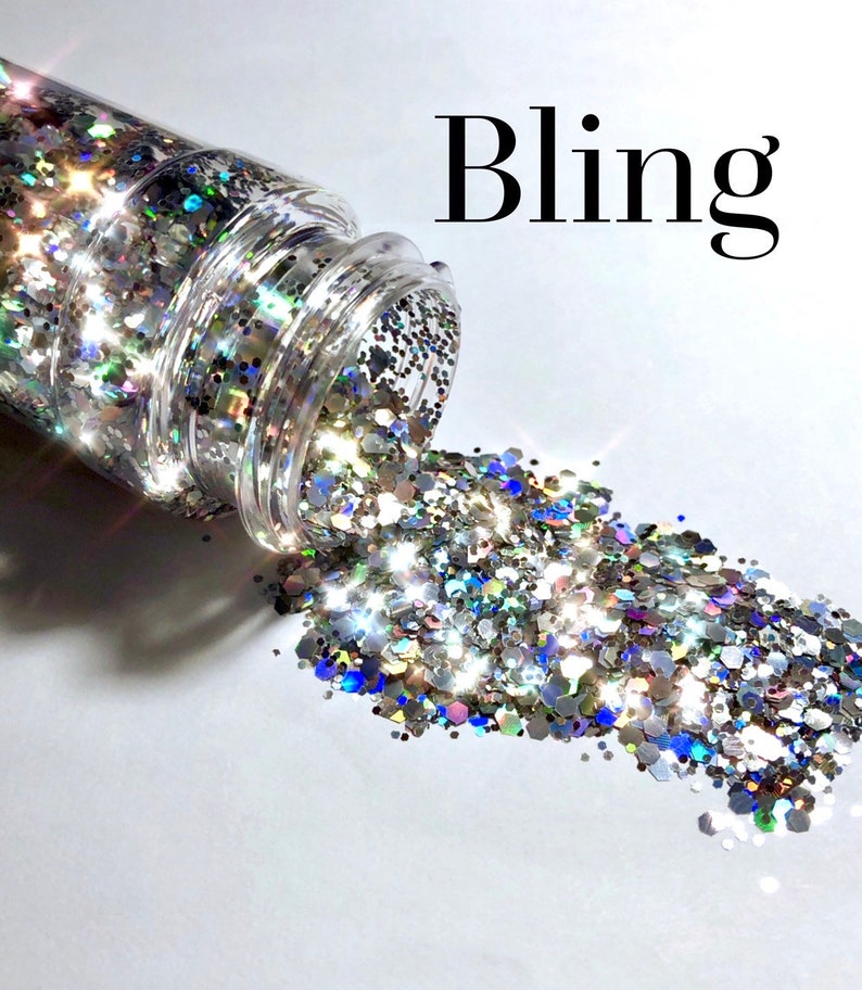Op de afbeelding: Een doorzichtige plastic pot met zilveren, blauwe en iriserende glitter die eruit stroomt. De glitter heeft verschillende vormen en maten. Het woord "Bling" staat in zwarte tekst boven de pot.