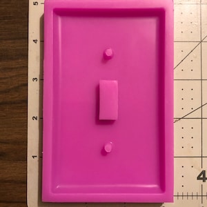 May include: A pink silicone mold for making a light switch cover. The mold has a rectangular shape with a raised outline of a light switch, including a toggle switch and two screw holes.