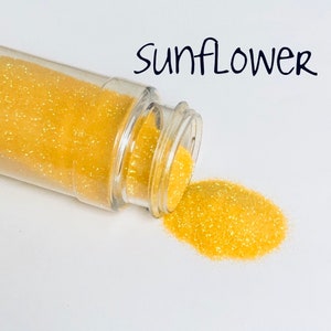 May include: A clear plastic bottle tipped over, spilling out a pile of yellow glitter. The glitter is labeled "sunflower" in black script. The glitter is a bright, shimmering yellow.