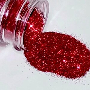 May include: A small jar of red glitter is spilling out onto a white surface. The glitter is fine and sparkly, and it creates a beautiful, shimmering effect.