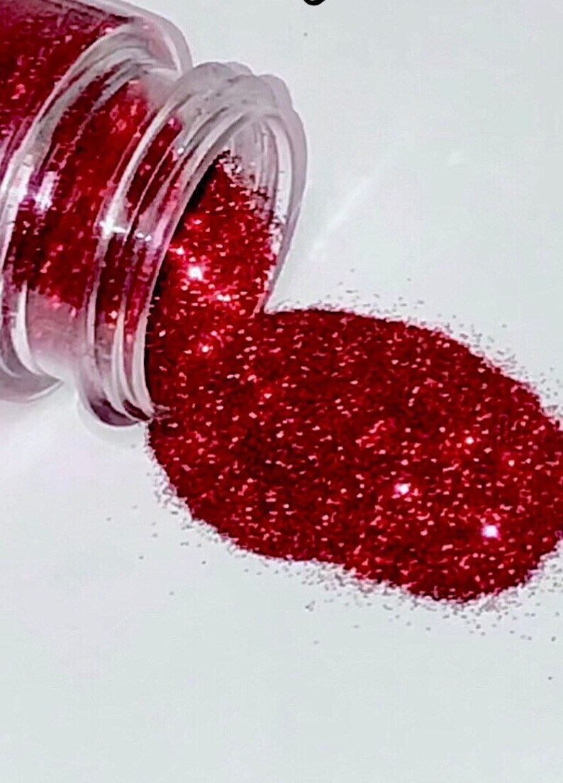 May include: A small jar of red glitter is spilling out onto a white surface. The glitter is fine and sparkly, and it creates a beautiful, shimmering effect.