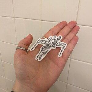 May include: A black and white line drawing of a Star Wars X-Wing fighter on a white background.