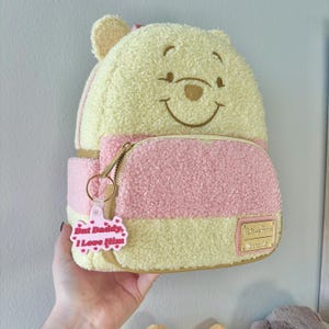 May include: A Winnie the Pooh-themed mini backpack in yellow and pink, featuring a smiling Pooh face on the front. The backpack has a front pocket, a gold zipper, and a pink keychain that says "But Daddy, I Love Him."