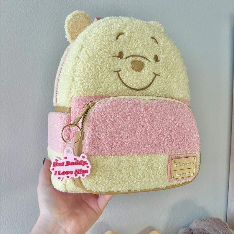 May include: A Winnie the Pooh-themed mini backpack in yellow and pink, featuring a smiling Pooh face on the front. The backpack has a front pocket, a gold zipper, and a pink keychain that says "But Daddy, I Love Him."