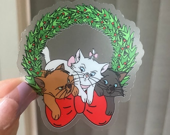 May include: Three cartoon cats wearing a red bow, surrounded by a green wreath with red berries. The cats are white, brown, and black. The white cat is in the center and has a pink bow on its head. The brown cat is on the left and the black cat is on the right.