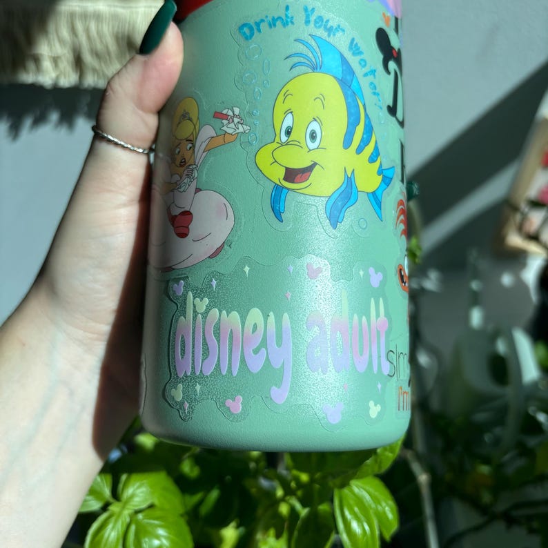 Op de afbeelding: Een lichtgroene waterfles met stickers van Disney-personages, waaronder Botje en Alice in Wonderland. De fles heeft de woorden "Drink Your Water" en "disney adults" erop gedrukt.