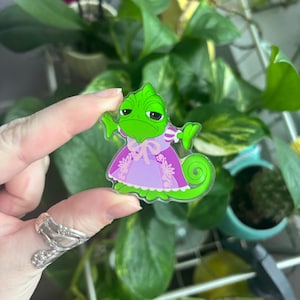 Puede incluir: Un pin de personaje de camaleón verde que lleva un vestido morado con detalles en rosa. El pin se sostiene frente a un fondo de plantas verdes. El camaleón tiene una expresión de mal humor.