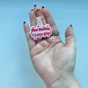 May include: A pink keychain with the phrase "But Daddy, I Love Him" in red lettering. The keychain has a gold-colored ring and a small, clear plastic attachment. The design includes small red heart accents.