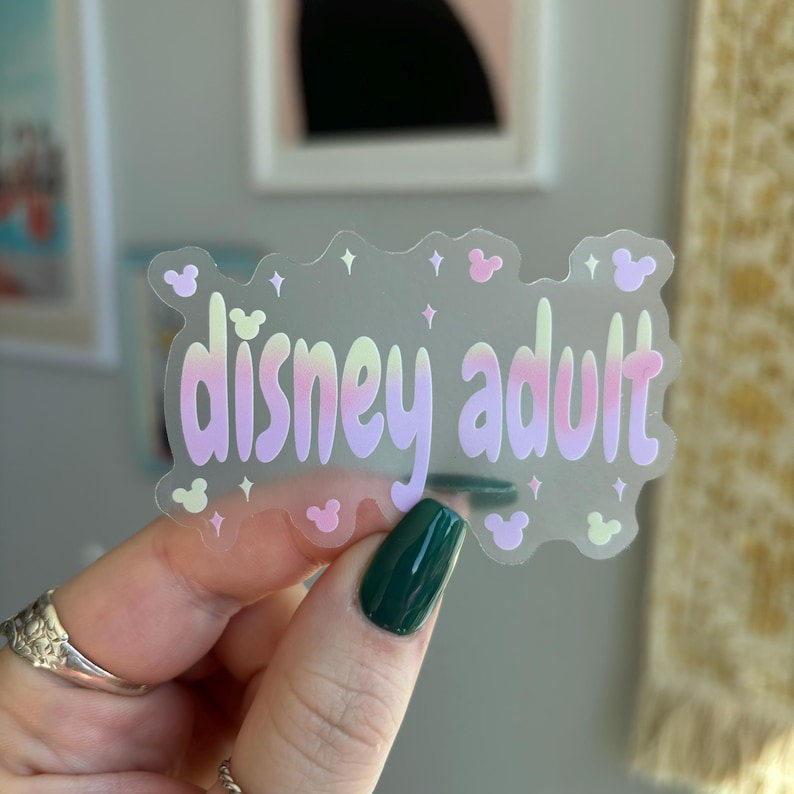 Op de afbeelding: Een doorzichtige sticker met de woorden "disney adult" in een pastelkleurige gradi&euml;ntlettertype. De sticker heeft een golvende omtrek met kleine Mickey Mouse-kop- en stervormen. De sticker wordt vastgehouden door een hand met donkergroene nagellak.