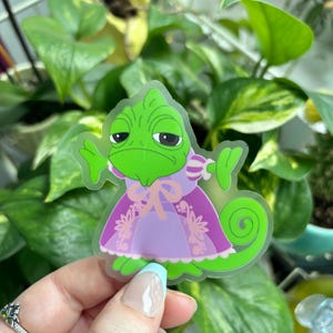 May include: A clear sticker of a green chameleon wearing a purple dress with pink floral details. The chameleon has a grumpy expression and is holding its arms out.