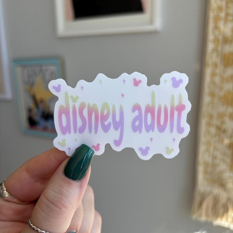 Op de afbeelding: Een witte sticker met de woorden "disney adult" in een verloop van roze, geel en paars. De sticker heeft een golvende omtrek met kleine Mickey Mouse-vormen en sterren. De sticker wordt vastgehouden door een hand met een groen gelakte nagel.