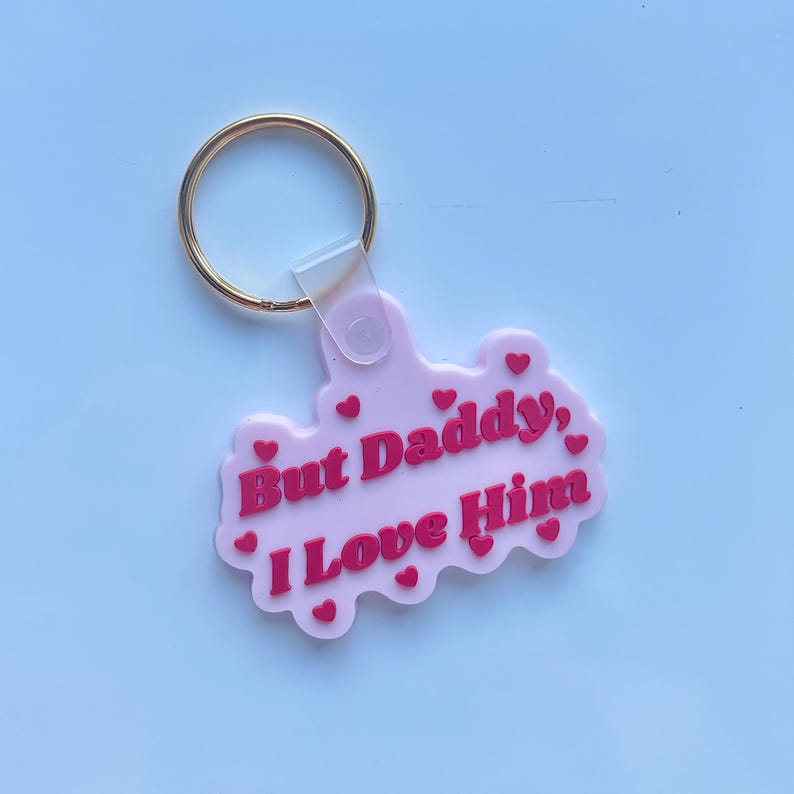May include: A pink keychain with a gold ring. The keychain has the phrase "But Daddy, I Love Him" in red lettering, with small red hearts scattered around the text. The keychain is made of a soft, flexible material.
