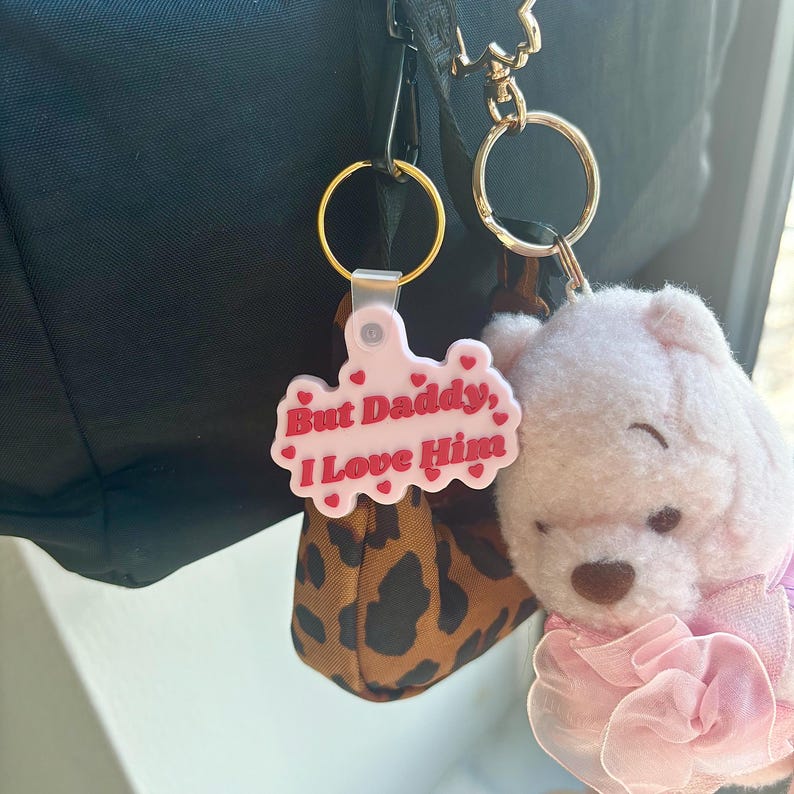 May include: A pink keychain with the text "But Daddy, I Love Him" in red, heart-shaped accents. A small, plush pink bear with a pink bow and a leopard print pouch are attached to the keychain.