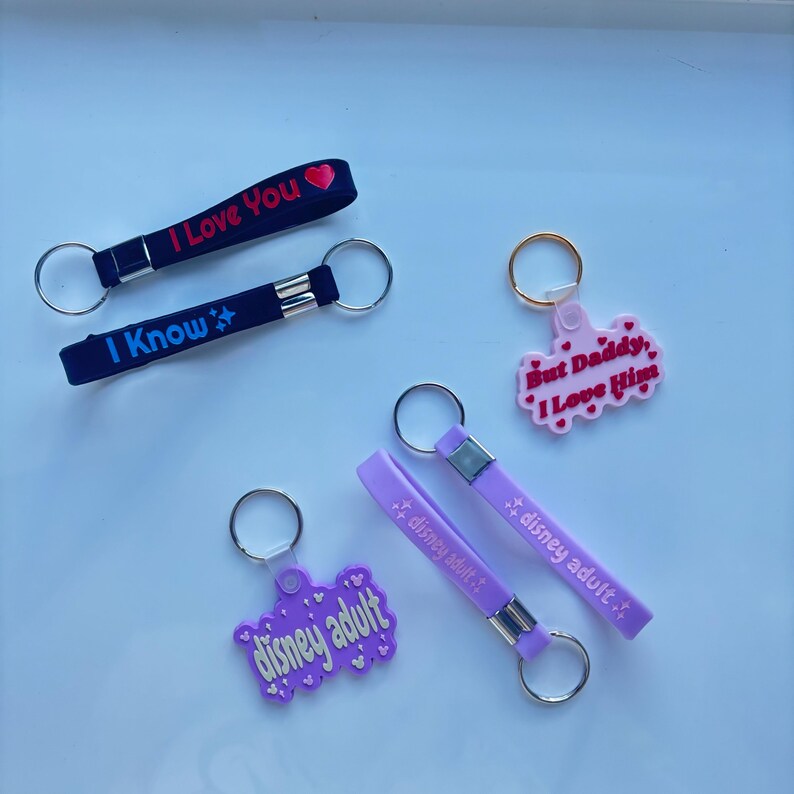 May include: Collection of keychains in various colors. Two black keychains with the phrases "I Love You" and "I Know". A pink keychain with "But Daddy, I Love Him". Two purple keychains with "disney adult". All have silver key rings.