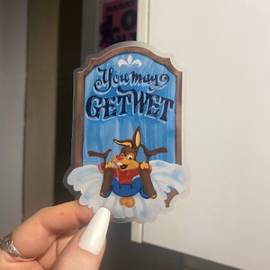 Puede incluir: Un cartel azul y blanco con el texto "You may get wet" en una fuente caprichosa. Un conejo de dibujos animados con una camisa roja y pantalones marrones está montando un tronco por una colina nevada.