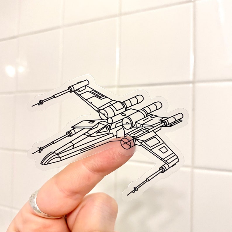 May include: A clear sticker of a black and white line drawing of an X-Wing starfighter from Star Wars.