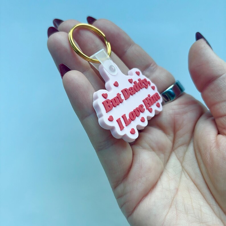May include: A pink keychain with the phrase "But Daddy, I Love Him" in red lettering. The keychain has a gold-colored ring and is decorated with small red hearts.