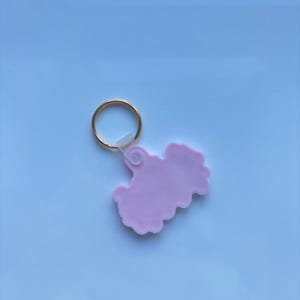 May include: A light pink keychain with a gold-colored ring. The keychain has a unique, cloud-like shape. The background is a light blue color.