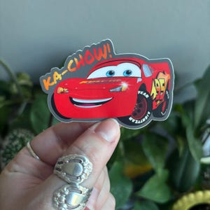 May include: A sticker featuring Lightning McQueen from the movie Cars. The red race car has a smiling face and the text "KA-CHOW!" above it. The number "95" is on the side. The sticker is on a gray background.