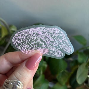 Può includere: Un adesivo trasparente con un contorno rosa dell'astronave Millennium Falcon. L'adesivo presenta un design dettagliato dell'esterno della nave, con vari pannelli e forme. L'adesivo è tenuto contro uno sfondo di fogliame verde.