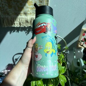 Op de afbeelding: Een lichtgroene waterfles met een zwarte dop, versierd met stickers van tekenfilmfiguren. De stickers bevatten een rode raceauto, een geel-blauwe vis en een prinses. De tekst "Drink Your Water" en "disney adults" is ook zichtbaar.