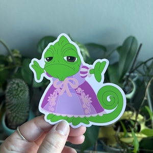Puede incluir: Una pegatina de camaleón verde con un vestido morado y rosa. El camaleón tiene una expresión aburrida y una cola enroscada. La pegatina se sostiene frente a un fondo de plantas verdes.