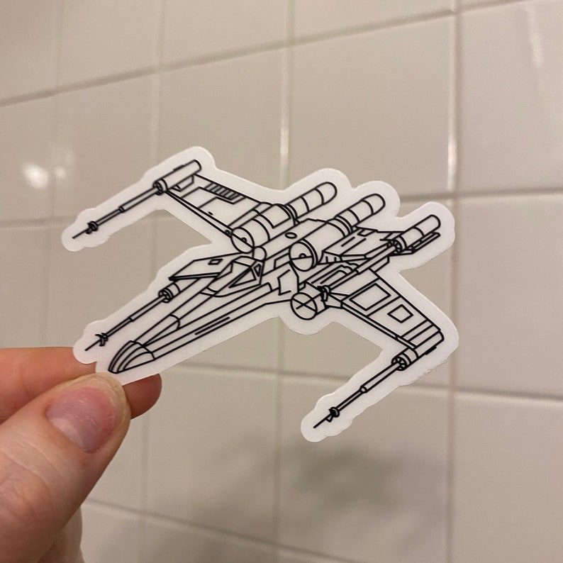 May include: A white outline sticker of an X-Wing starfighter from Star Wars. The sticker is on a white tiled background.
