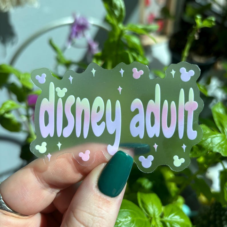 Op de afbeelding: Een transparante sticker met de woorden "disney adult" in een pastelkleurige gradi&euml;ntlettertype. De sticker heeft een lichtgroene achtergrond met roze Mickey Mouse-vormen en steraccenten. De sticker wordt voor een achtergrond van groene planten gehouden.