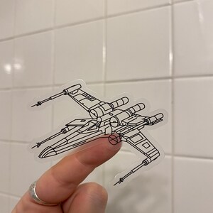 May include: A clear sticker with a black line drawing of an X-Wing starfighter from Star Wars. The sticker is being held by a finger.