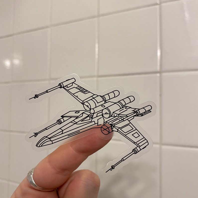 May include: A clear sticker with a black line drawing of an X-Wing starfighter from Star Wars. The sticker is being held by a finger.
