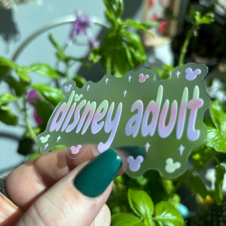Op de afbeelding: Een transparante sticker met de woorden "disney adult" in een grillige lettertype. De sticker heeft een lichtgroene achtergrond met roze en paarse accenten en kleine Mickey Mouse-hoofdvormen. De sticker wordt voor een achtergrond van groene planten gehouden.