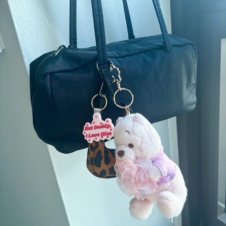 May include: Black nylon handbag with two attached keychains. One keychain is a leopard print design with the text "But Daddy, I Love Him". The other is a plush teddy bear wearing a pink outfit.