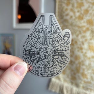 Könnte beinhalten: Ein transparenter Aufkleber mit einer schwarzen Linienzeichnung des Raumschiffs Millennium Falcon. Die Zeichnung zeigt die Details des Innen- und Außendesigns des Schiffes.