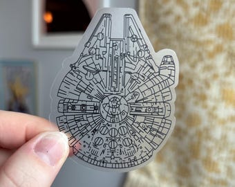 Może przedstawiać: Przezroczysta naklejka z czarnym rysunkiem linii statku kosmicznego Millennium Falcon. Rysunek przedstawia szczegóły wnętrza i zewnętrza statku.