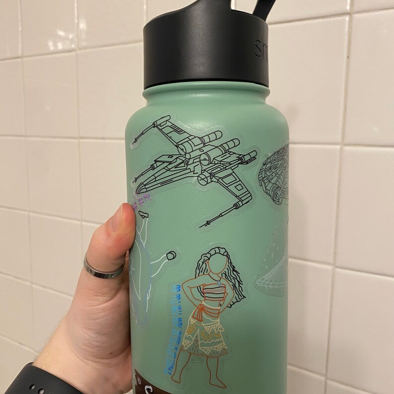 May include: A green reusable water bottle with a black lid and a variety of stickers. The stickers include a cartoon of a spaceship, a cartoon of a woman in a dress, and a cartoon of a woman with a flower in her hair.
