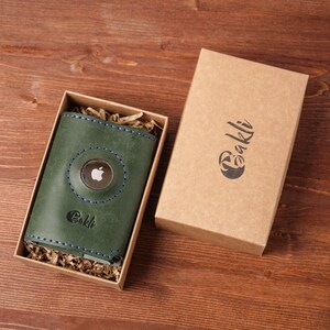 May include: A green leather wallet with blue stitching and an Apple logo, presented in a kraft paper gift box. The wallet has the word "Bakli" embossed on it. The box also has the word "Bakli" printed on it.