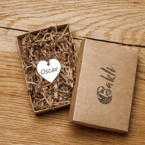 May include: A brown cardboard box with the word "Bakli" printed on it. The box is open and contains a small silver heart-shaped charm with the name "Oscar" engraved on it.