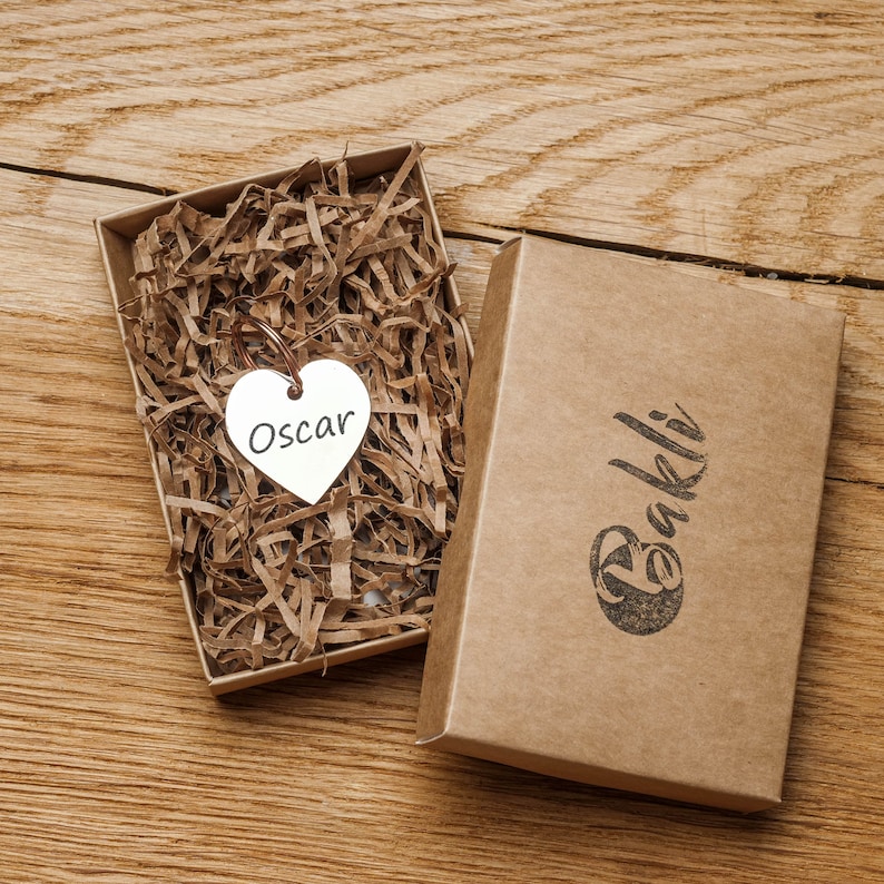 May include: A brown cardboard box with the word "Bakli" printed on it. The box is open and contains a small silver heart-shaped charm with the name "Oscar" engraved on it.