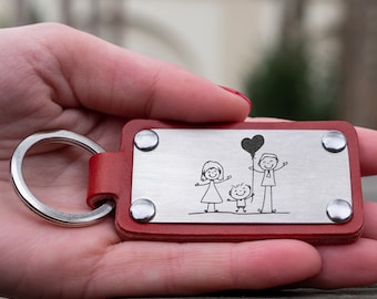 Puede incluir: Llavero de cuero rojo con un rectángulo plateado que presenta una ilustración dibujada a mano de una familia con un globo en forma de corazón. El llavero tiene un anillo plateado.