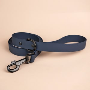Op de afbeelding: Een donkerblauwe rubberen hondenriem met een zwarte metalen clip en een zwarte metalen ring. De riem is opgerold en klaar voor gebruik.