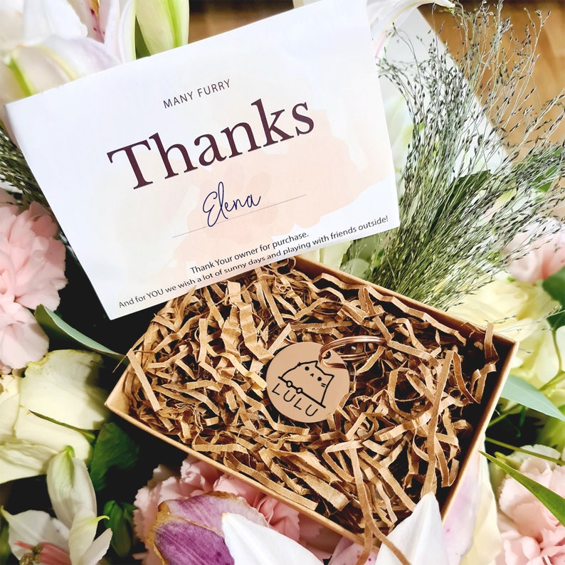 Op de afbeelding: Een bruine cadeaudoos gevuld met bruin geribbeld papier en een koperkleurig kattenvormig hondenpenning met de naam "LULU" erop gegraveerd. De doos is omringd door roze en witte bloemen. Een kaart met de tekst "MANY FURRY Thanks Elena Thank Your owner for purchase. And for YOU we wish a lot of sunny days and playing with friends outside!" is zichtbaar op de achtergrond.