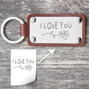 Könnte beinhalten: Ein brauner Leder-Schlüsselanhänger mit einem silbernen Metalletikett, auf dem "I LOVE YOU" in handschriftlicher Schrift mit einem einfachen Blumendesign steht.