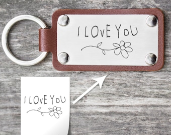 Puede incluir: Un llavero de cuero marrón con una etiqueta de metal plateado que dice "I LOVE YOU" en letra manuscrita con un diseño de flor simple.