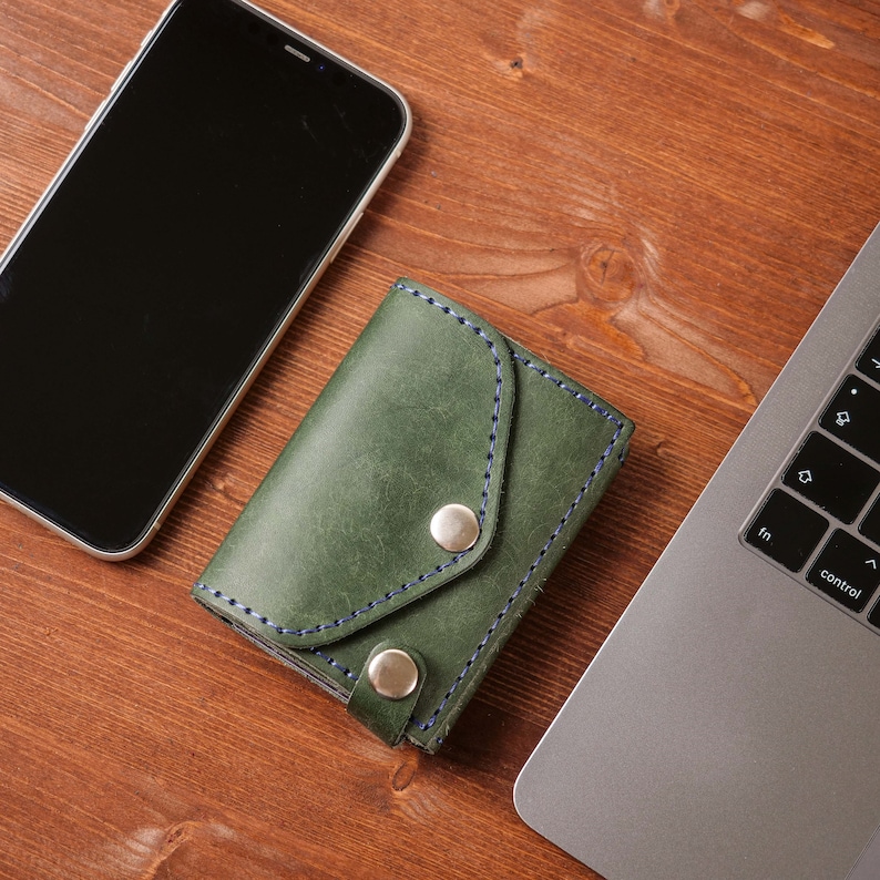 May include: A green leather card holder with blue stitching and two silver snap closures. The card holder is on a wooden surface next to a smartphone and a laptop.