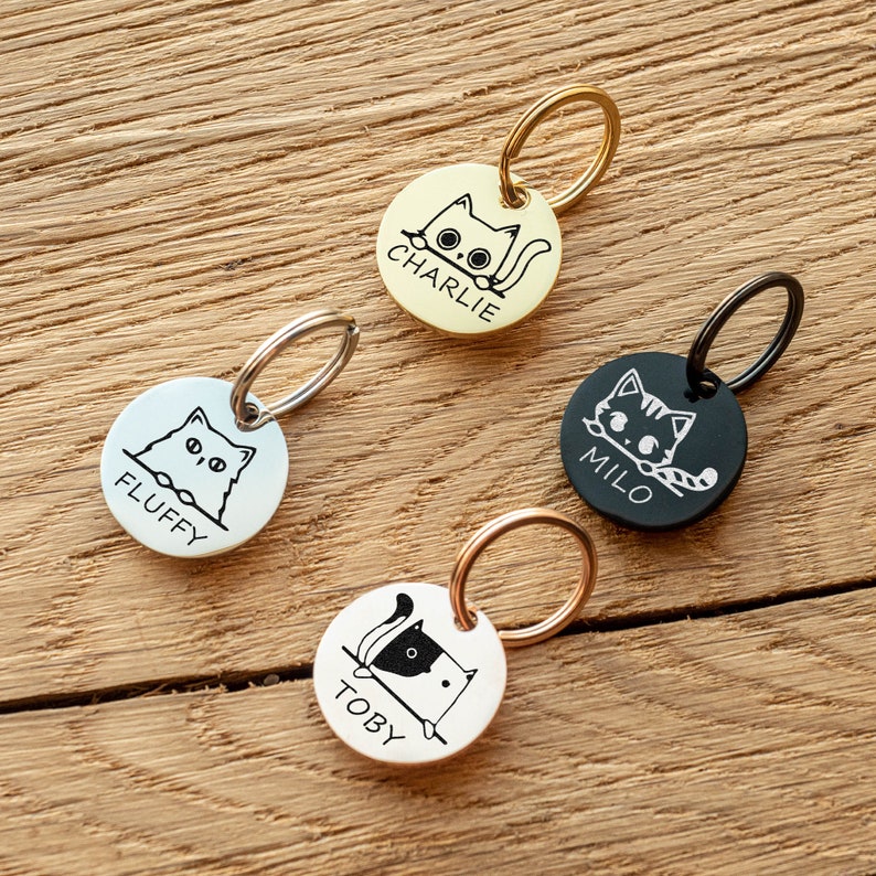 Op de afbeelding: Vier ronde katten-thema huisdier ID-tags met verschillende namen gegraveerd. De tags zijn zilver, goud, zwart en ros&eacute; goud. Elke tag heeft een kleine ring eraan bevestigd.