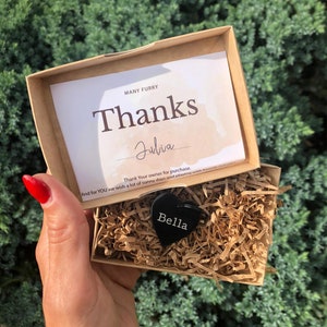 May include: A small, black heart-shaped dog tag with the name "Bella" engraved on it. The tag is in a brown cardboard box with shredded paper inside. A handwritten note in the box says "Many Furry Thanks Julia Thank Your owner for purchase. And for YOU we wish a lot of sunny days and playing with friends."