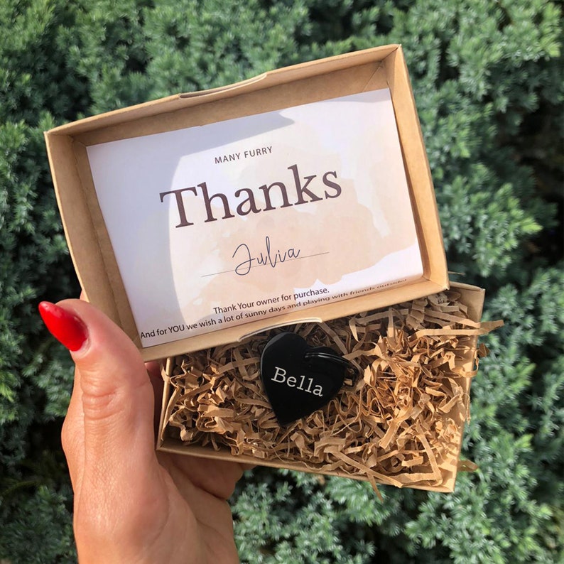 May include: A small, black heart-shaped dog tag with the name "Bella" engraved on it. The tag is in a brown cardboard box with shredded paper inside. A handwritten note in the box says "Many Furry Thanks Julia Thank Your owner for purchase. And for YOU we wish a lot of sunny days and playing with friends."