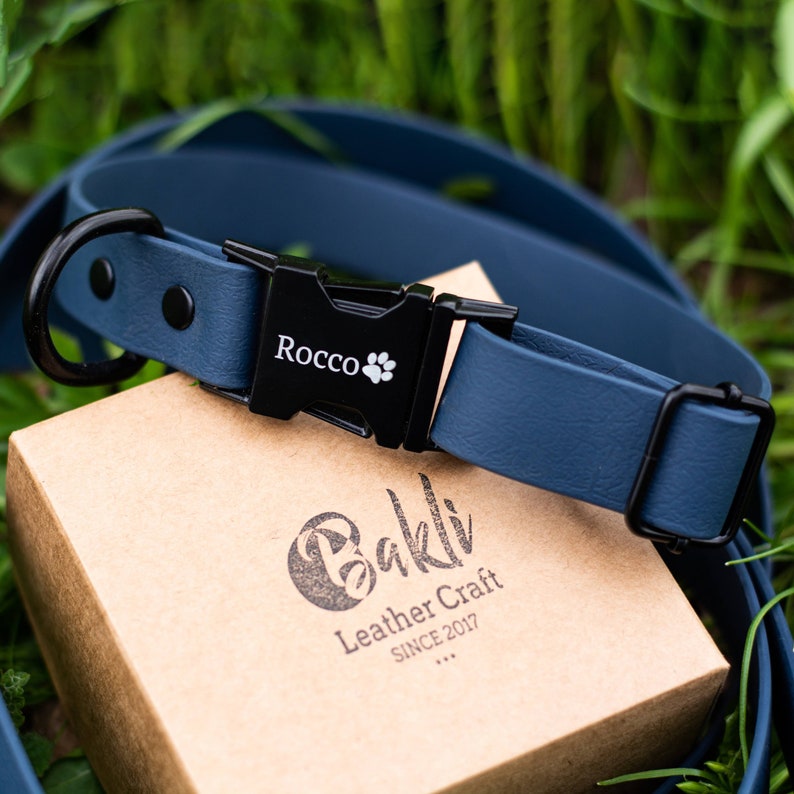 May include: A blue dog collar with a black buckle and a black D-ring. The buckle is engraved with the name "Rocco" and a paw print. The collar is on a brown box with the text "Bakli Leather Craft SINCE 2017" printed on it.