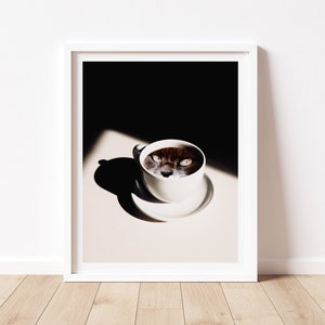May include: A white coffee cup with a cat's face peering out of the coffee. The cat's eyes are wide open and staring directly at the viewer. The cup is sitting on a white saucer on a light brown surface. The background is dark.
