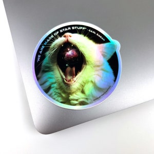 May include: A holographic sticker featuring a white cat with its mouth open, revealing a galaxy inside. The sticker has the text "We are made of star stuff" and "Carl Sagan" around the cat's head.